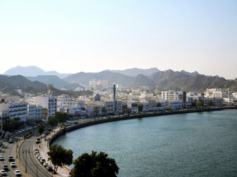 Aerial view of Muscat's white low-rise waterfront buildings along the Muttrah coastline, backed by rugged mountains and the Arabian Sea in Oman