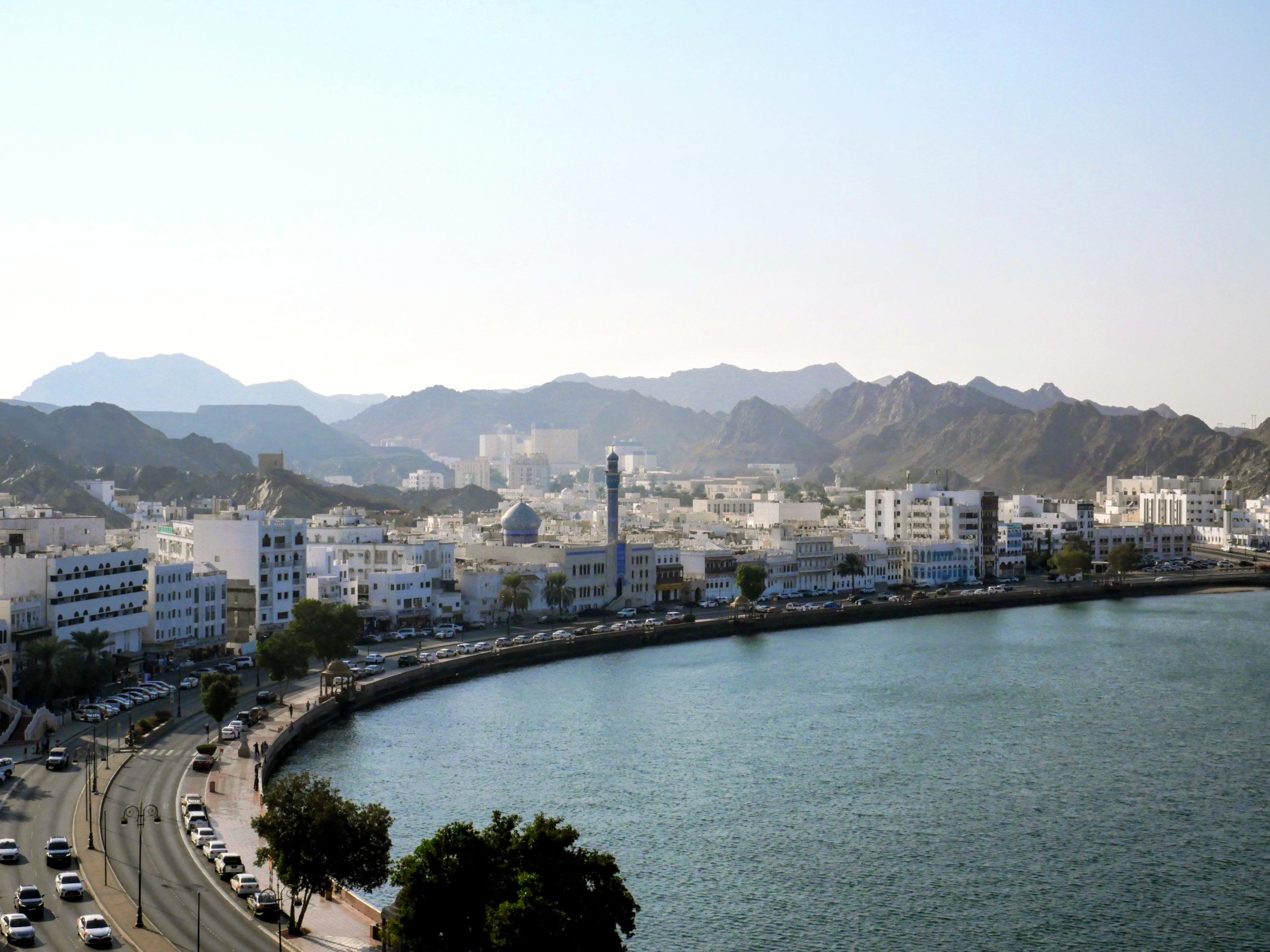 Aerial view of Muscat's white low-rise waterfront buildings along the Muttrah coastline, backed by rugged mountains and the Arabian Sea in Oman