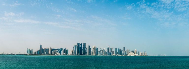 Wide daytime panorama of the Doha, Qatar city skyline, with the modern West Bay skyscrapers rising above the waterfront under a clear sky, conveying a prosperous, liveable Gulf capital for relocating expats and families.