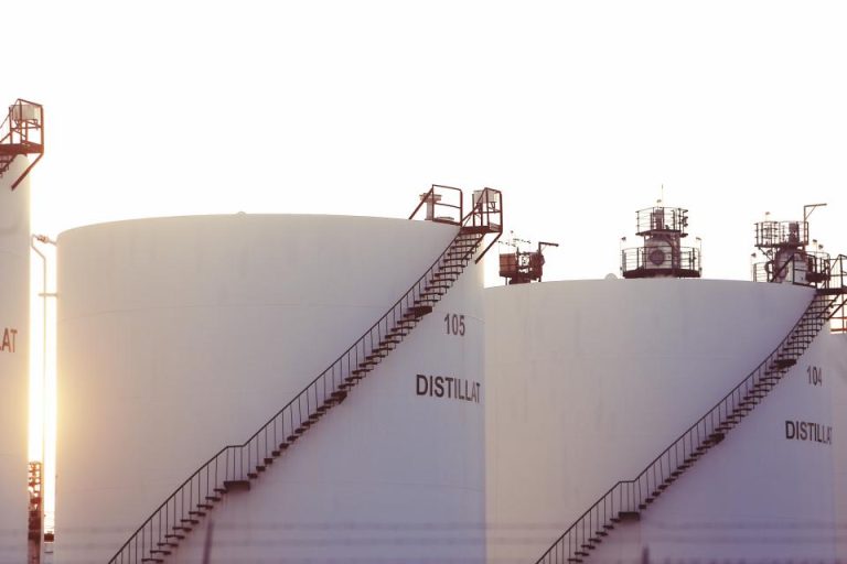 Rows of large white distillate storage tanks with access stairways at an oil and gas processing plant glowing in twilight sunlight, evoking a high-stakes LNG plant commissioning push.