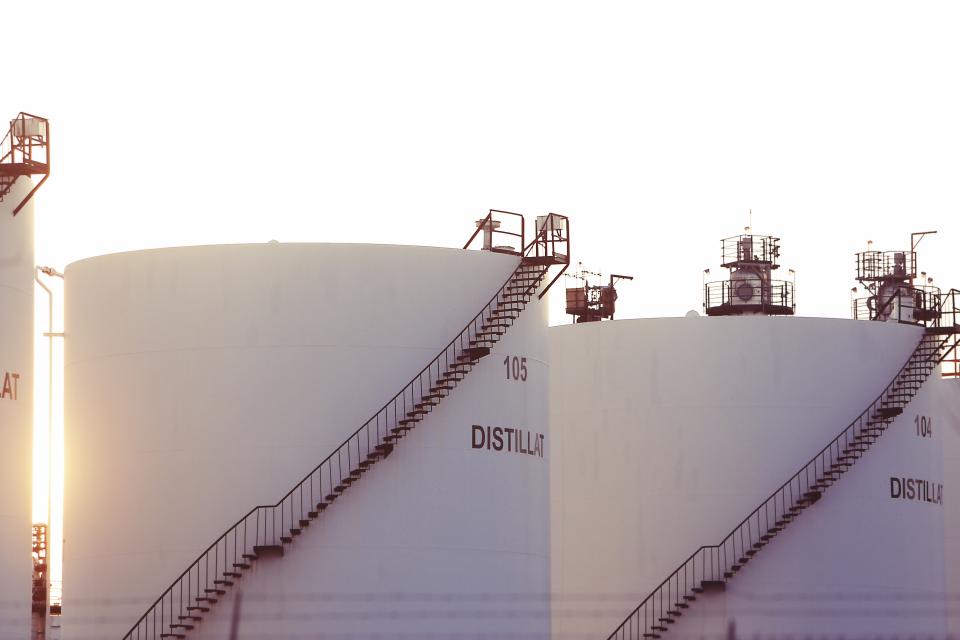 Rows of large white distillate storage tanks with access stairways at an oil and gas processing plant glowing in twilight sunlight, evoking a high-stakes LNG plant commissioning push.